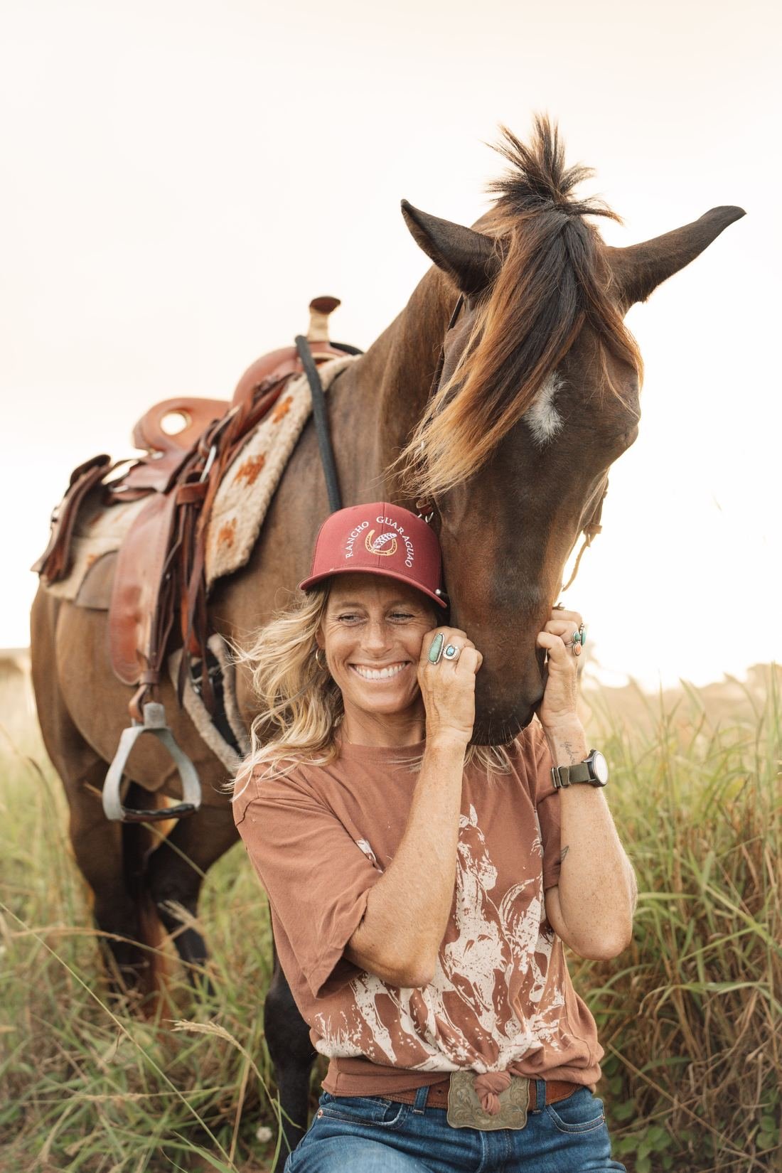 Smiling rider in a Rancho Guaraguao cap hugging her bay horse in a tall-grass field in Isabela, Puerto Rico